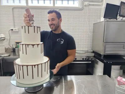 Pasticcere al lavoro su una drip cake bianca a tre piani, mentre applica la colatura di cioccolato lungo il bordo superiore. L'immagine valorizza la cura artigianale della pasticceria firmata Viglianisi Catering.