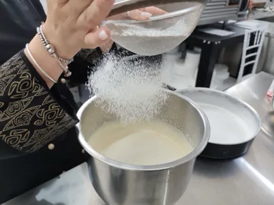 Preparazione di pasticceria con ingredienti secchi setacciati dentro una grande ciotola di impasto chiaro. La foto racconta un passaggio tecnico del laboratorio dolci di Viglianisi Catering.