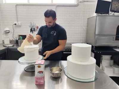Pasticcere al lavoro con sac à poche su una torta in fase di montaggio, mentre sul banco accanto si vedono altre basi già rivestite di bianco. L'immagine documenta una fase avanzata della lavorazione artigianale in pasticceria.