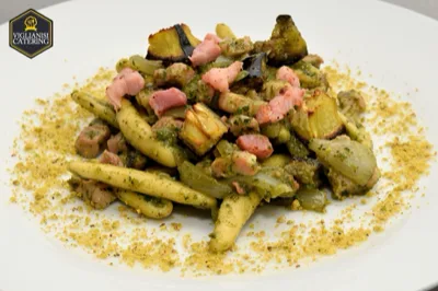 Piatto di pasta corta con condimento verde, cubetti rosa di salume o pesce e verdure saltate, rifinito con una spolverata croccante distribuita sul bordo del piatto. L'impiattamento mette in evidenza una proposta di primo piatto articolata e ricca di contrasti.