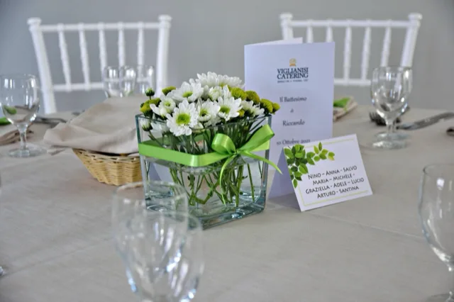 Centrotavola per battesimo con piccoli fiori bianchi raccolti in un contenitore di vetro trasparente legato da un nastro verde. Accanto si vedono il menu o programma dell'evento, il segnaposto degli invitati e una mise en place sobria con sedie bianche.