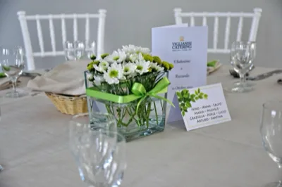 Centrotavola per battesimo con piccoli fiori bianchi raccolti in un contenitore di vetro trasparente legato da un nastro verde. Accanto si vedono il menu o programma dell'evento, il segnaposto degli invitati e una mise en place sobria con sedie bianche.