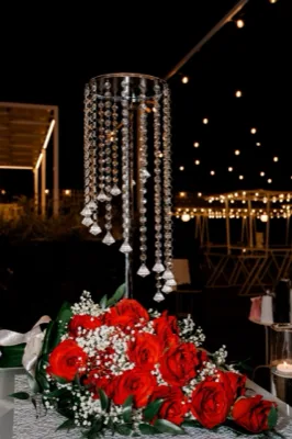 Centrotavola scenografico con rose rosse, gypsophila e foglie verdi alla base di un supporto alto decorato con pendenti di cristallo. Lo sfondo notturno con luci calde sospese enfatizza l'effetto elegante e teatrale dell'allestimento.