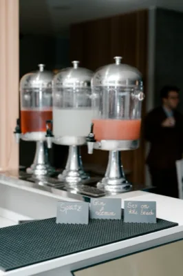 Tre dispenser di cocktail serviti su banco bar, con bevande di colori diversi e cartoncini con i nomi dei drink in primo piano. La postazione è ordinata e pensata per il servizio self-service durante l'evento.