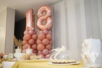 Composizione per i 18 anni con numero 18 in palloncini color oro rosa, colonne di palloncini rosa e allestimento da festa in un ambiente interno elegante. Sul tavolo davanti si intravedono decorazioni e vassoi di dolci.