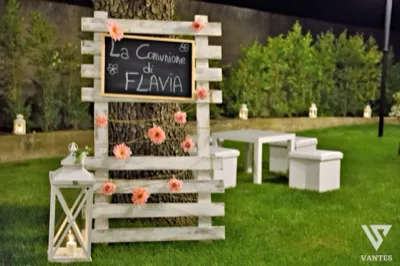 Allestimento esterno serale per una Comunione con lavagna decorativa fissata a una struttura di legno chiaro, arricchita da piccoli fiori rosa. In primo piano si vede una lanterna bianca accesa sul prato, mentre sullo sfondo compaiono sedute bianche e il giardino illuminato.