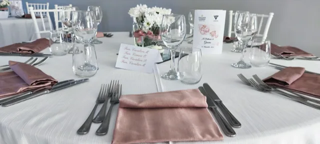 Dettaglio di un tavolo da battesimo apparecchiato con tovagliato bianco, tovaglioli rosa cipria, calici trasparenti e cartoncino centrale con il nome degli invitati. Al centro c'è anche un piccolo vaso con fiori bianchi, per una mise en place sobria e raffinata.