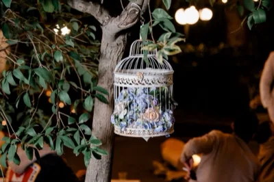 Gabbia decorativa bianca appesa a un albero nel giardino, riempita di fiori azzurri e rosa con fogliame verde. Lo sfondo notturno è illuminato da luci calde e la scena richiama un allestimento romantico per evento all'aperto.