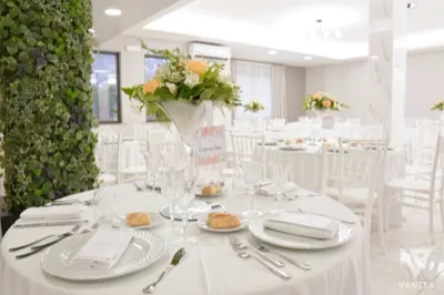 Tavolo rotondo apparecchiato in una sala ricevimenti luminosa, con piatti bianchi, calici lavorati e centrotavola alto con composizione floreale chiara. Sullo sfondo si vedono altri tavoli coordinati e l'ambiente elegante dai toni neutri.