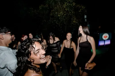 Gruppo di ragazzi che balla e ride durante una festa all'aperto di notte, con la postazione DJ visibile sullo sfondo. La scena è vivace e illuminata da luce flash e da piccoli punti luce caldi tra gli alberi.