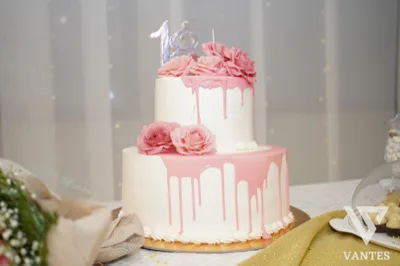Torta drip a due piani per i 18 anni, decorata con colature rosa, rose in tonalità rosa e numero 18 in cima. La torta è impostata su un tavolo elegante con sfondo chiaro e luci soffuse.