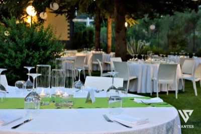 Cena all'aperto con tavoli apparecchiati, tovagliato bianco, runner verde e calici trasparenti sotto alberi illuminati. Sullo sfondo si vedono altre tavolate e l'atmosfera rilassata di un ricevimento serale.