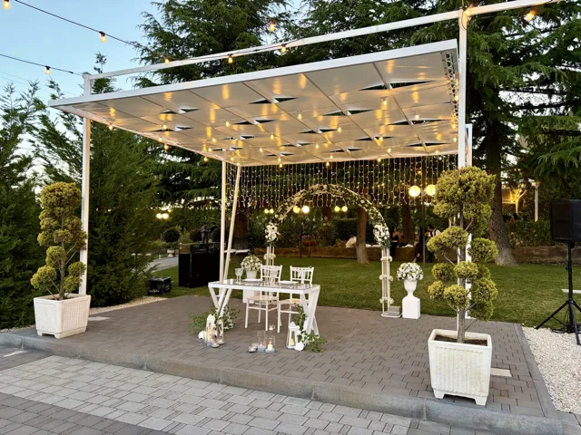 Gazebo esterno illuminato con tavolo cerimonia bianco, arco floreale sullo sfondo e lanterne decorative ai piedi della struttura. Le catenarie e il prato serale creano un'ambientazione elegante per il rito o il taglio torta del matrimonio.