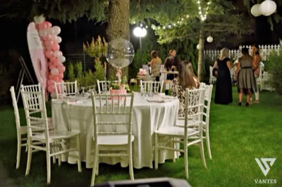 Tavolo rotondo apparecchiato sul prato durante una serata in giardino, con sedie bianche, centrotavola e ospiti distribuiti tra gli alberi illuminati. Sul lato sinistro si nota anche una scenografia con palloncini rosa che contribuisce all'atmosfera della festa.