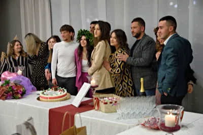 Gruppo di ragazzi e adulti raccolti attorno al tavolo torta durante una festa di laurea in sala, con la laureata al centro che indossa corona d'alloro e tailleur fucsia. In primo piano si vedono la torta decorata, un bouquet di fiori viola, calici allineati e candele accese che completano l'allestimento del brindisi.