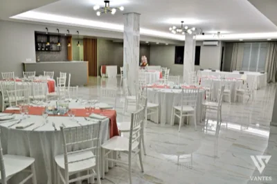 Sala ricevimenti interna con tavoli rotondi apparecchiati, sedie bianche, runner rosa salmone e pavimento lucido effetto marmo. Sullo sfondo si vedono il bancone bar, i pilastri chiari e un'impostazione ordinata adatta a un ricevimento elegante.