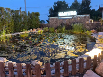 Laghetto decorativo al crepuscolo con ninfee, canneti, rocce e riflessi luminosi sull'acqua davanti a una parete in pietra. Sullo sfondo spicca l'insegna Vantes illuminata sopra la cascata, elemento scenografico distintivo della location.