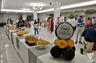 Festa privata in sala con lungo buffet salato disposto su più tavoli, ospiti in piedi e personale di servizio distribuito nell'ambiente. In primo piano si nota una decorazione con palloncini neri e oro e scritta Happy Birthday che identifica il contesto celebrativo.