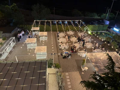 Vista aerea notturna della location con tavoli rotondi, buffet e aree relax distribuite nel cortile. Le luci sospese e il laghetto illuminato evidenziano la geometria dell'allestimento dall'alto.