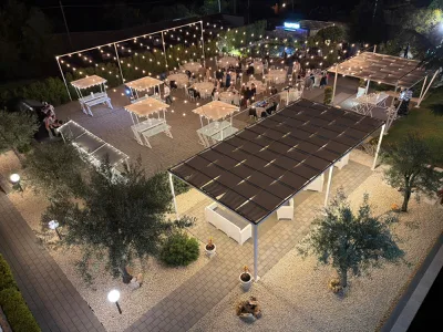 Vista alta notturna della location con tavoli rotondi illuminati, buffet laterali e fili di luci sospese che attraversano l'intero spazio. Al centro si vede la distribuzione ordinata degli ospiti e, sul fondo, il laghetto decorativo.