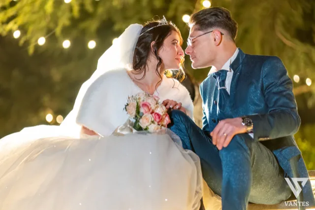 Ritratto notturno ravvicinato degli sposi seduti sotto luci calde in giardino, mentre si guardano sorridendo con bouquet rosa e abito da cerimonia in evidenza. L'atmosfera è intima e romantica, con sfondo sfocato e illuminazione soffusa.