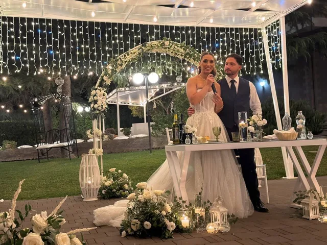 Sposa con microfono accanto allo sposo sotto un arco floreale, davanti a un tavolo allestito con candele, bottiglie e piccoli vasi bianchi. Le luci pendenti e il prato sullo sfondo definiscono una scena serale da discorso o brindisi durante il matrimonio.