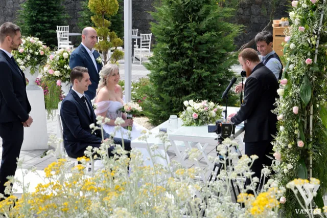 Momento del rito civile all'aperto con celebrante al tavolo, sposi seduti davanti alle composizioni floreali e testimoni ai lati. L'inquadratura diurna mostra il set della cerimonia tra fiori chiari, prato e dettagli decorativi.