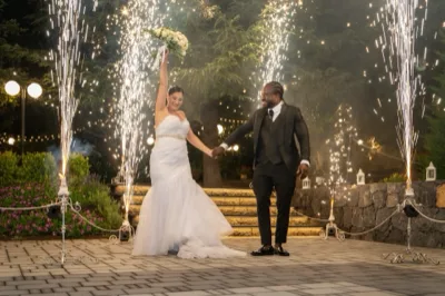 Sposi in abiti da cerimonia che avanzano mano nella mano all'aperto di sera, circondati da fontane luminose e scintille scenografiche. La scena mette in risalto l'ingresso spettacolare del ricevimento, con gradini illuminati e giardino sullo sfondo.