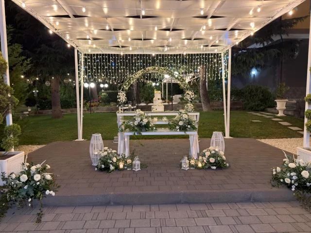Allestimento serale per matrimonio con tavolo bianco, torta a piani al centro e grande arco floreale sotto il gazebo illuminato. Le lanterne, le composizioni di rose bianche e il prato sullo sfondo costruiscono una scenografia elegante per il taglio torta.