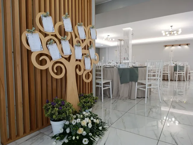Tableau mariage a forma di albero in legno con cartoncini numerati e fiocchi verde salvia, posizionato all'ingresso della sala. Sullo sfondo compaiono tavoli rotondi apparecchiati, pavimento lucido e centrotavola pendenti coordinati.
