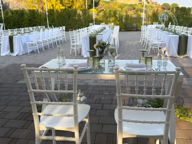 Tavolo degli sposi in primo piano davanti a lunghe tavolate imperiali allestite all'esterno, con sedie bianche, candele in cilindri di vetro e dettagli verdi coordinati. L'immagine valorizza la disposizione simmetrica della cena di matrimonio nel giardino.