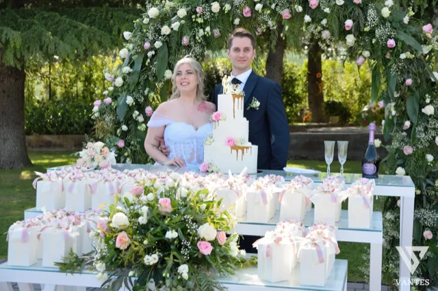 Sposi in posa dietro il tavolo torta in giardino, incorniciati da un arco floreale ricco di rose bianche e rosa. In primo piano si vedono bomboniere coordinate, composizioni floreali e una torta bianca a tre piani con dettagli dorati.