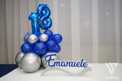 Composizione per i 18 anni in blu e argento con numeri metallici in cima e nome Emanuele in primo piano. La base è formata da palloncini di varie dimensioni e la scena è pensata come decorazione da tavolo o ingresso.