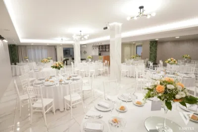 Panoramica della sala Vantes allestita con tavoli rotondi bianchi, sedie coordinate e composizioni floreali chiare come centrotavola. Lo spazio appare luminoso, ordinato e rifinito con pavimento effetto marmo, pilastri chiari e bancone bar sul fondo.