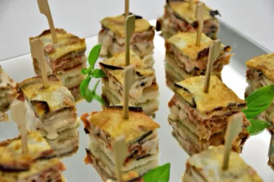 Piccoli tranci di finger food stratificato con melanzane, formaggio fuso e pane, serviti con stecchino su un vassoio a specchio. Il primo piano evidenzia la consistenza filante del ripieno e la presentazione curata da buffet.