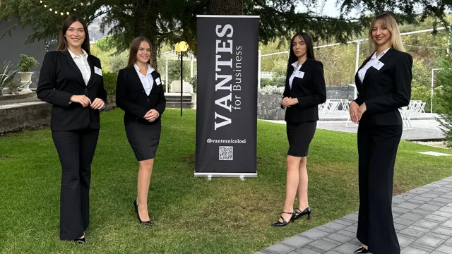 Quattro persone dello staff in divisa elegante posano sul prato accanto a un banner Vantes for Business. Sullo sfondo si vedono il giardino della location, luci sospese e l'area esterna allestita per eventi aziendali.