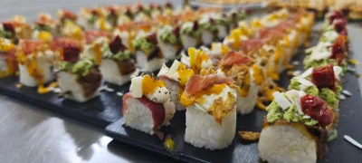 Primo piano ravvicinato di rolls di sushi creativi disposti su vassoi neri, con topping colorati, creme e ingredienti decorativi. L'immagine mette in evidenza la varietà e la presentazione del buffet catering.