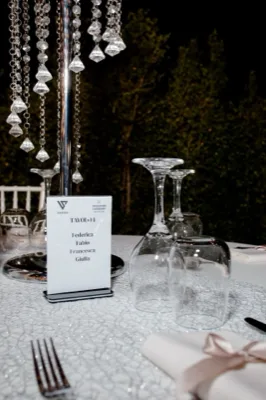 Dettaglio di un tavolo apparecchiato di sera con segnatavolo nominativo, calici capovolti e centrotavola con pendenti in cristallo. La mise en place è elegante e curata, con tovagliato chiaro e sfondo scuro del giardino.