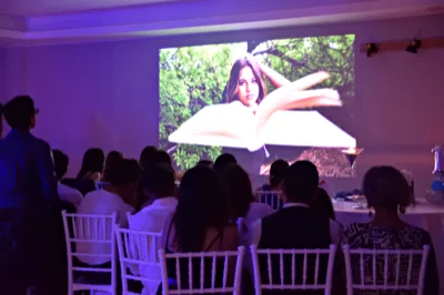 Ospiti seduti in sala durante la proiezione di un video o slideshow su una grande parete, in un ambiente illuminato da luci soffuse violacee. La scena racconta un momento raccolto e celebrativo all'interno della festa.