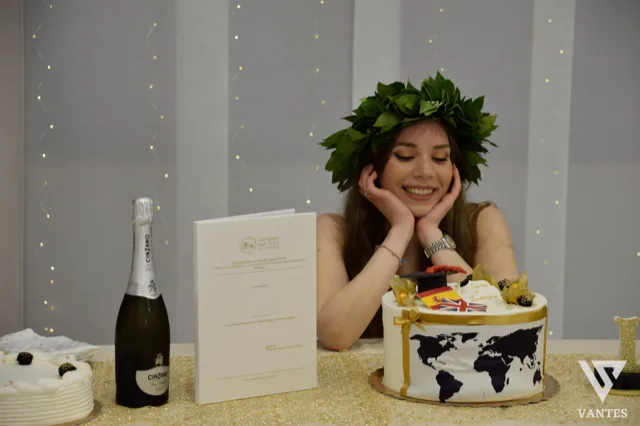 Laureata sorridente con corona d'alloro seduta dietro il tavolo torta, mentre appoggia il viso sulle mani in una posa spontanea. Accanto a lei si vedono una bottiglia di spumante, la tesi universitaria in verticale, una torta bianca decorata con mappamondo nero e dettagli oro e un allestimento luminoso sullo sfondo.