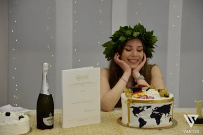Laureata sorridente con corona d'alloro seduta dietro il tavolo torta, mentre appoggia il viso sulle mani in una posa spontanea. Accanto a lei si vedono una bottiglia di spumante, la tesi universitaria in verticale, una torta bianca decorata con mappamondo nero e dettagli oro e un allestimento luminoso sullo sfondo.