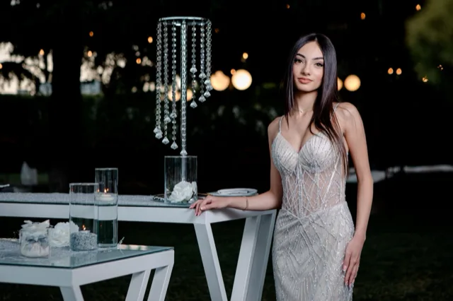 Ritratto serale di un'ospite accanto a tavoli alti decorati con candele, fiori bianchi e pendenti in cristallo. Le luci soffuse del giardino sullo sfondo rafforzano l'atmosfera elegante della location.