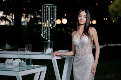 Ritratto serale di un'ospite accanto a tavoli alti decorati con candele, fiori bianchi e pendenti in cristallo. Le luci soffuse del giardino sullo sfondo rafforzano l'atmosfera elegante della location.