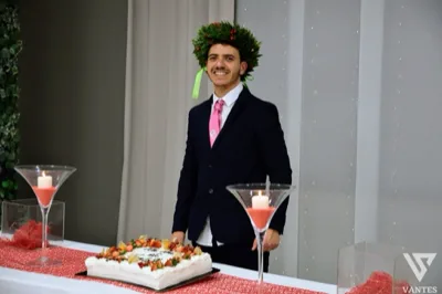 Ragazzo laureato in abito scuro con cravatta rosa e corona d'alloro, in posa sorridente dietro il tavolo torta durante il ricevimento. In primo piano compaiono una torta rettangolare decorata con fragole, due grandi candelieri con candele accese e dettagli rossi coordinati con l'allestimento.