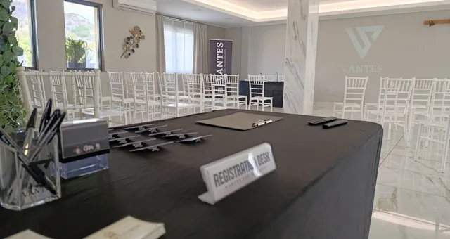 Desk registrazione per meeting allestito con penne, badge e cartello registration desk in primo piano, davanti a una sala con file di sedie bianche. Sullo sfondo si vedono il logo Vantes proiettato e un banner dedicato agli eventi business.