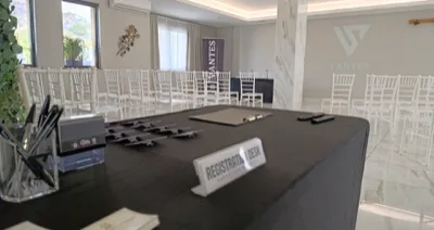 Desk registrazione per meeting allestito con penne, badge e cartello registration desk in primo piano, davanti a una sala con file di sedie bianche. Sullo sfondo si vedono il logo Vantes proiettato e un banner dedicato agli eventi business.