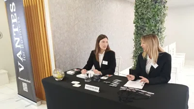Desk registrazione presidiato da due persone dello staff in divisa, con badge, blocchi appunti e materiale informativo per un evento business. Accanto al tavolo si vede il banner Vantes for Business e sullo sfondo l'ingresso della sala con le sedie già disposte.