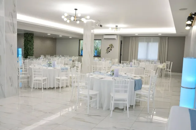 Sala interna allestita per un battesimo con tavoli rotondi bianchi, sedie coordinate, dettagli azzurri sui runner e piccoli centrotavola floreali. L'ambiente è molto luminoso e ordinato, con elementi decorativi cilindrici illuminati sui lati.