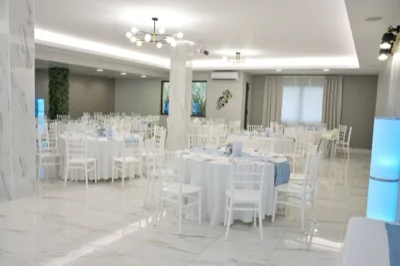 Sala interna allestita per un battesimo con tavoli rotondi bianchi, sedie coordinate, dettagli azzurri sui runner e piccoli centrotavola floreali. L'ambiente è molto luminoso e ordinato, con elementi decorativi cilindrici illuminati sui lati.