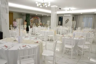 Sala interna apparecchiata per cerimonia con tavoli rotondi bianchi, sedie chiavari, runner chiari e alti centrotavola pendenti. Sullo sfondo si vede un arco di palloncini nei toni del rosa e dell'oro con tavolo decorativo coordinato.